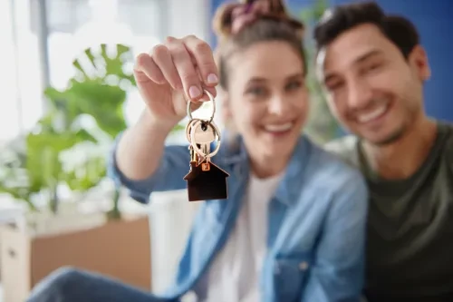 asal jovem sentado em casa, sorridente, enquanto a mulher segura um molho de chaves com um porta-chaves em forma de casa, simbolizando a compra da primeira habitação.
