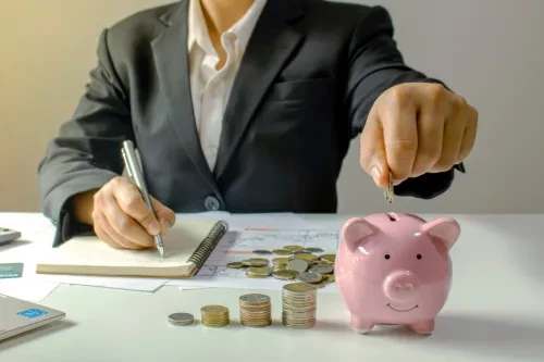 Pessoa de fato a registar informações financeiras enquanto coloca uma moeda num mealheiro cor-de-rosa, com várias pilhas de moedas ao lado, representando poupança e gestão financeira.