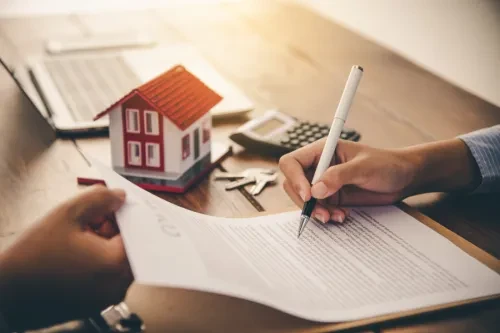 Duas pessoas a analisar e assinar documentação de crédito habitação, com uma miniatura de casa, chaves e calculadora sobre a mesa, simbolizando contratação ou renegociação de crédito.