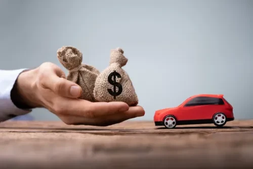 Mão a segurar sacos de dinheiro com símbolo de dólar enquanto um carro vermelho em miniatura está pousado à frente, representando financiamento automóvel.
